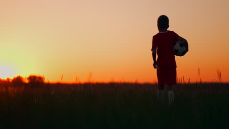 The-boy-goes-to-the-field-at-sunset-with-a-soccer-ball-looking-into-the-distance-and-dreams-of-becoming-a-successful-football-player.-Watch-the-sunset-in-the-field